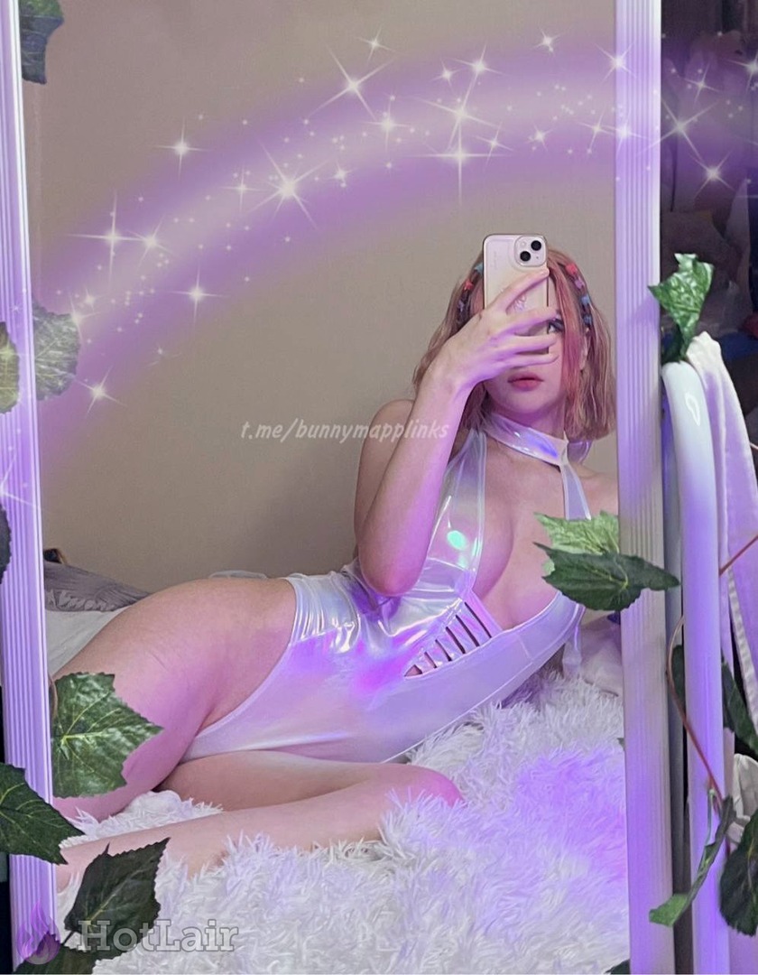 Stunning pink-haired beauty in shiny holographic bodysuit lying seductively on fluffy rug mirror selfie