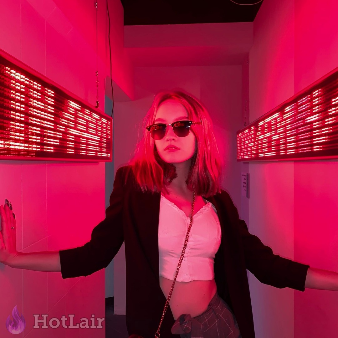 Seductive brunette posing in black blazer and white crop top under vibrant red neon lights with sunglasses