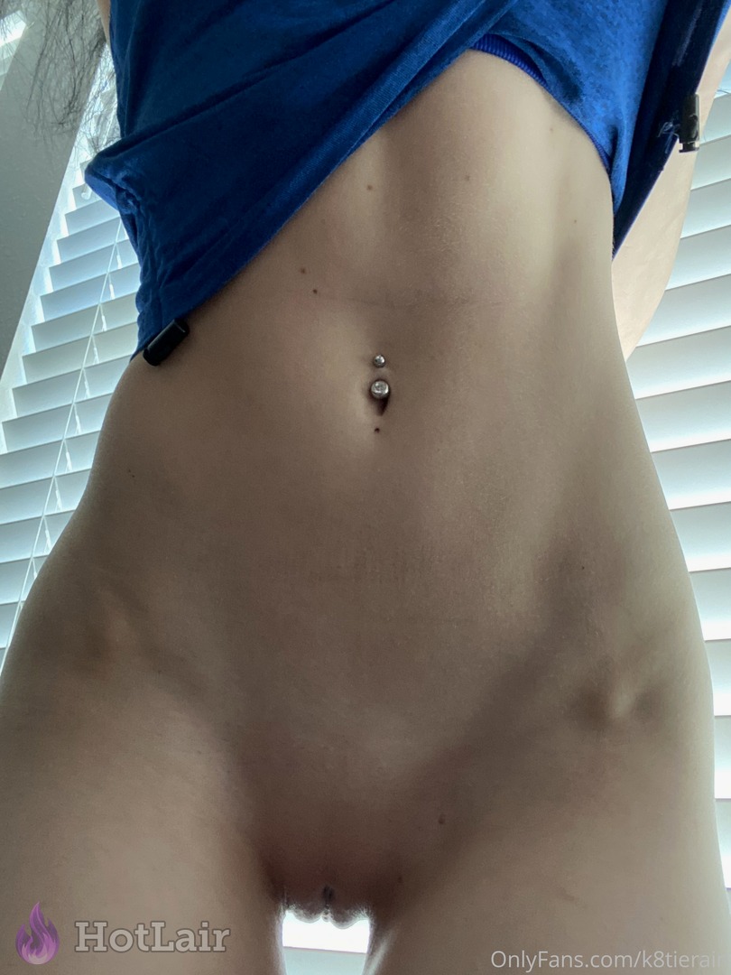 Close-up erotic shot of brunette’s toned stomach, navel piercing and shaved pussy under lifted blue top with natural light