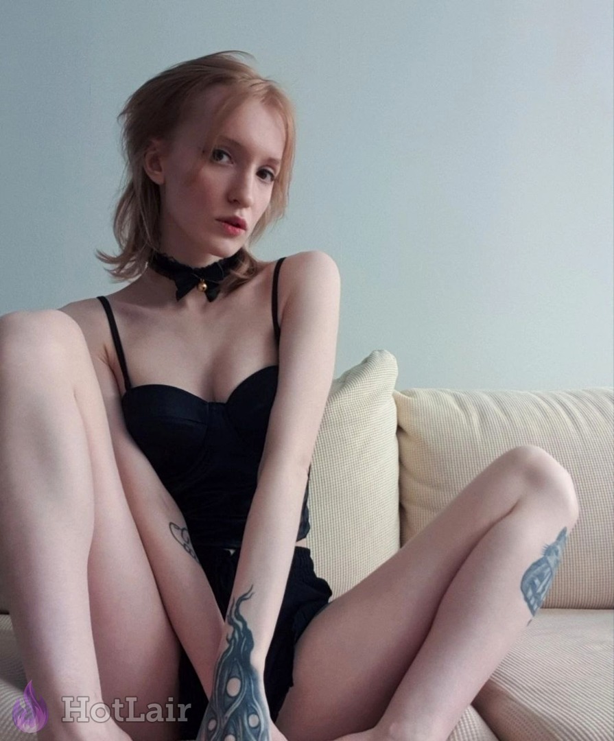Stunning redhead with tattoos and black choker sitting on sofa in sexy black lingerie, legs crossed and seductive stare at camera