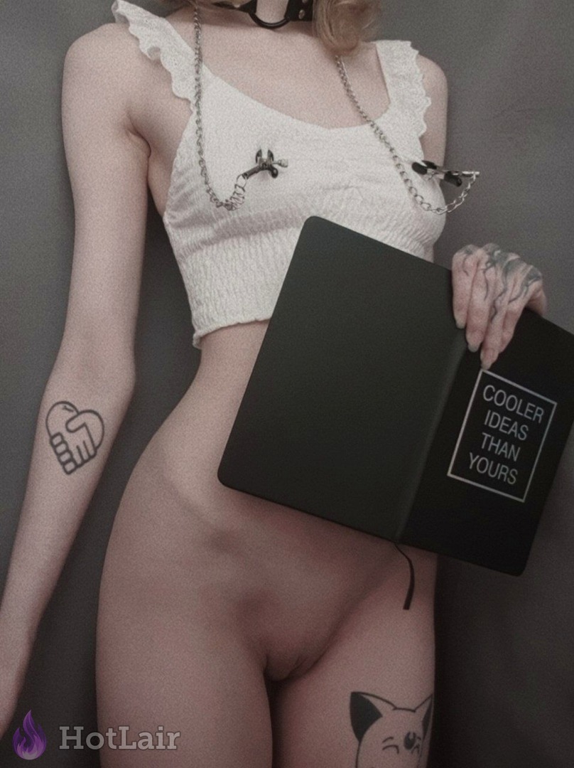 Topless tattooed woman in white ruffled crop top with nipple clamps and chains, holding black book in provocative pose