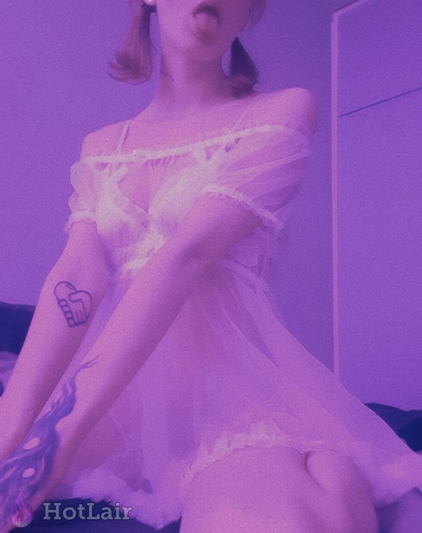 Cute blonde in sheer white off-shoulder lingerie dress sticking tongue out playfully, purple lighting and visible tattoos