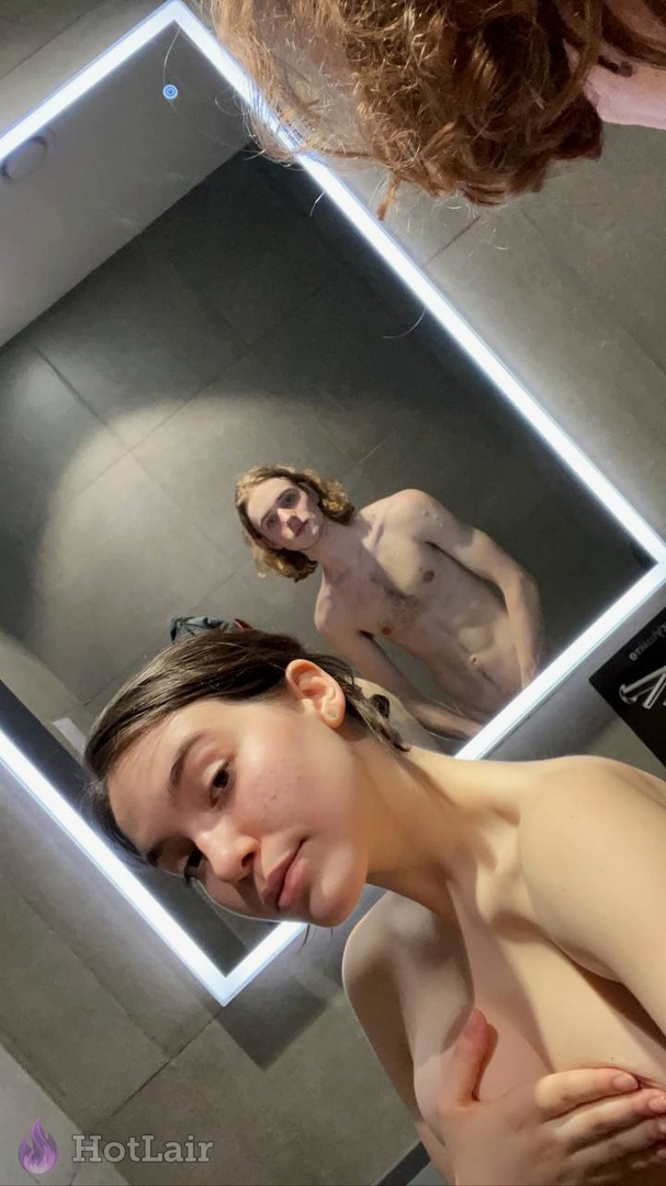 Topless girl taking mirror selfie with friend visible in reflection, hand covering breast seductively