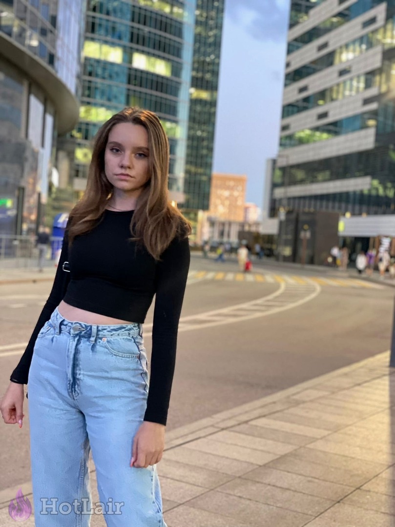 Pretty young brunette in black crop top and light blue jeans posing on city street with modern buildings in background