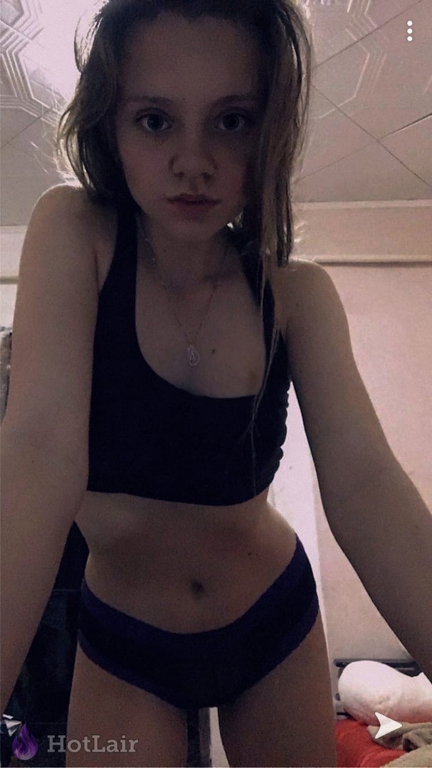 Slim young woman in black crop top and panties taking mirror selfie in bedroom, looking straight at camera