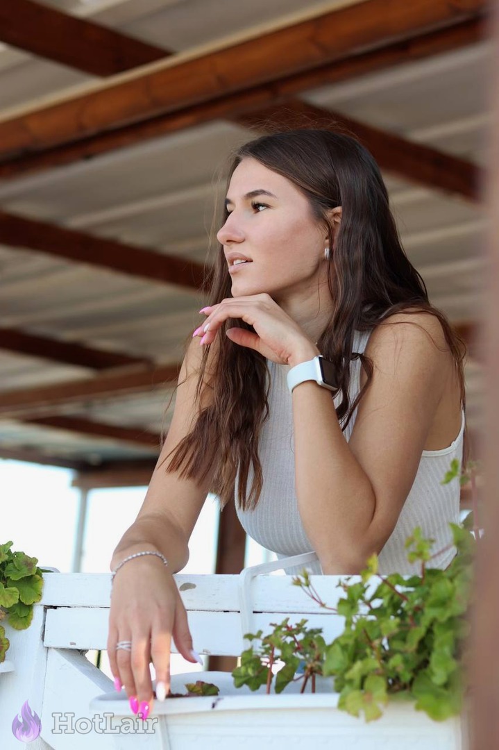 Beautiful long-haired brunette in white ribbed top posing thoughtfully on balcony with hand on chin