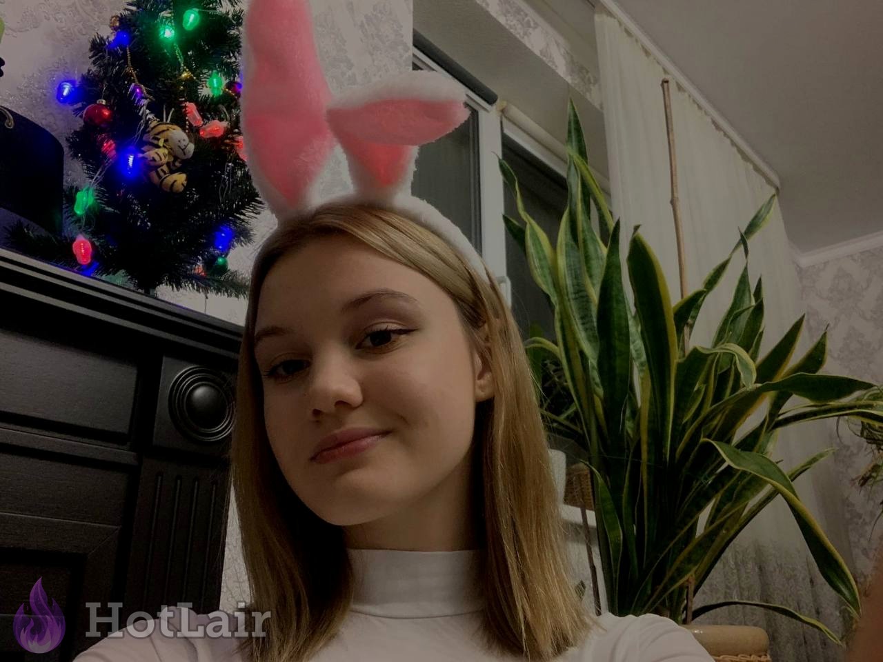 Adorable blonde girl wearing cute pink bunny ears headband smiling playfully near Christmas tree
