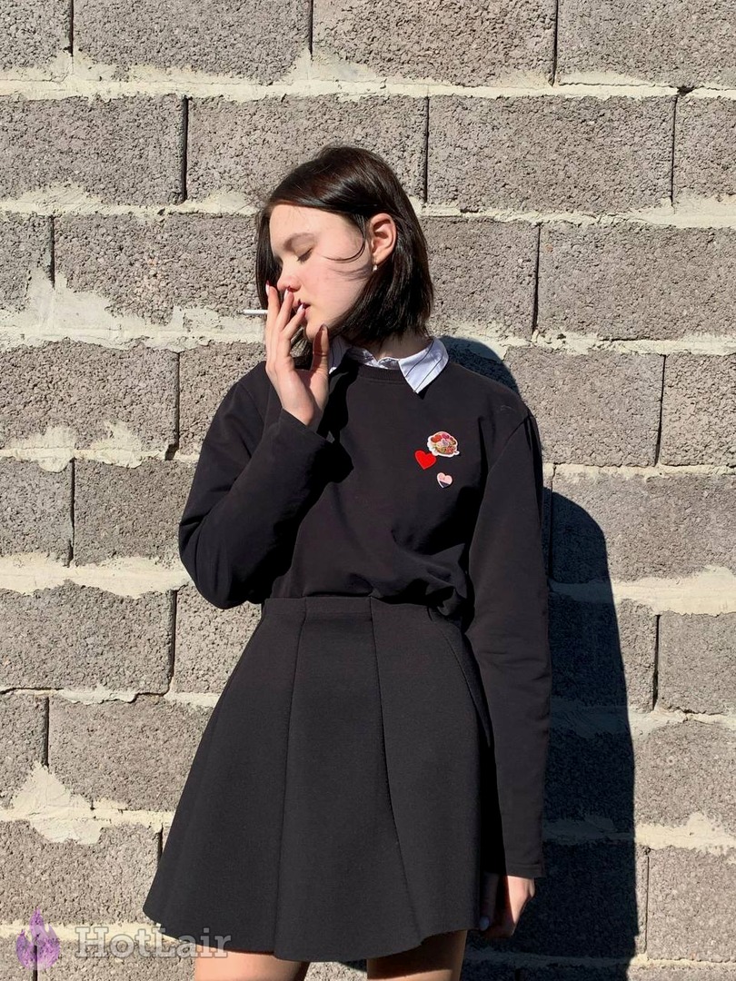 Stylish young brunette in black sweater and pleated skirt smoking against brick wall, casual outdoor portrait