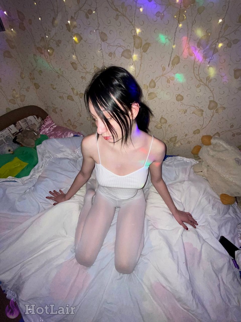 Sexy brunette in white crop top and sheer tights kneeling seductively on bed under colorful rainbow string lights