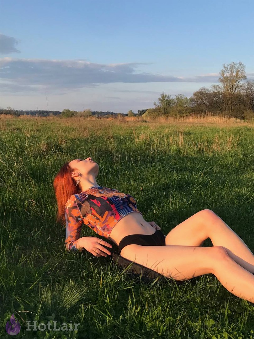 Gorgeous redhead lying topless in green field at sunset wearing colorful sheer top and black shorts
