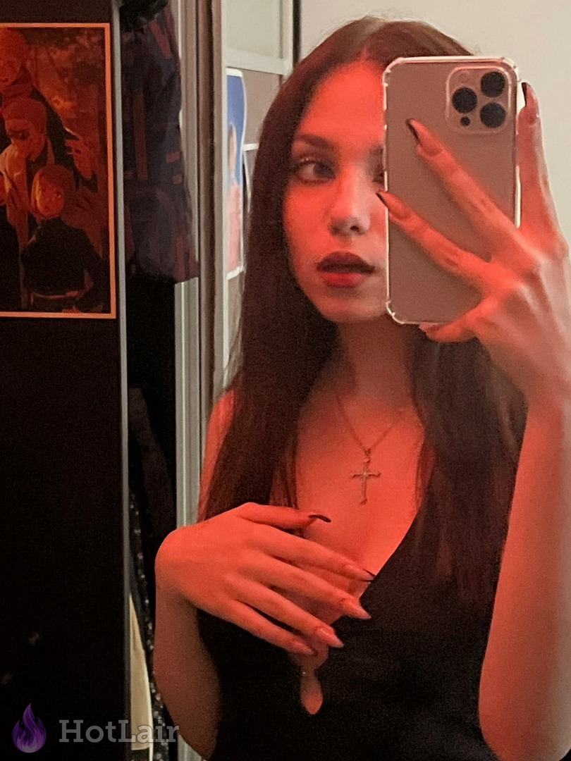 Stunning brunette with long dark hair and red lipstick taking mirror selfie in black dress under red lighting, seductive look
