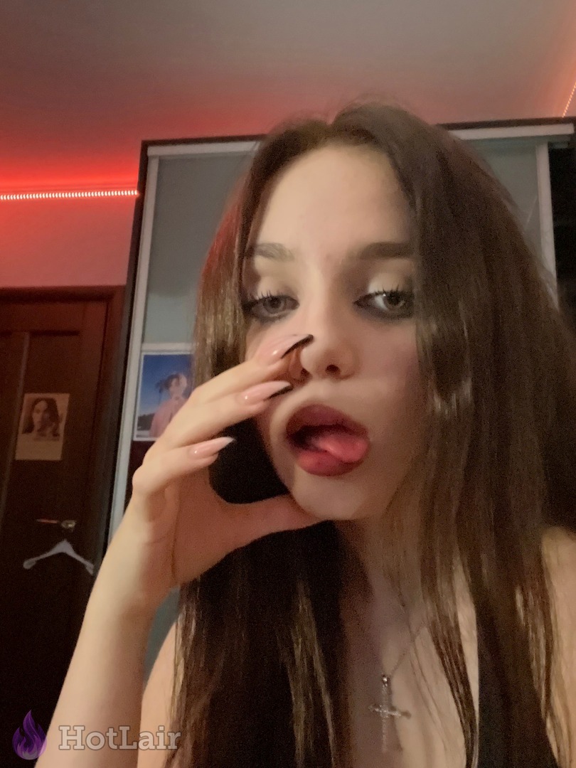 Playful brunette sticking out tongue and touching nose with long nails in close-up selfie with cross necklace