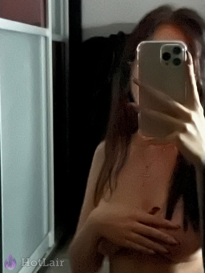 Topless brunette covering breasts with hand while taking mirror selfie in dim light, erotic and teasing