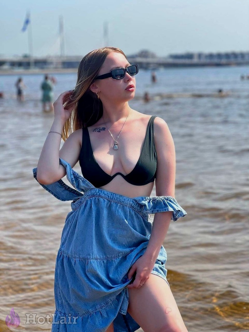 Seductive blonde in black bikini top and denim skirt posing by the sea, hand in hair with sunglasses and summer vibe