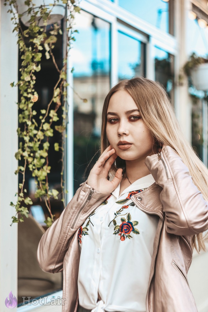 Beautiful long-haired blonde in pink leather jacket and embroidered shirt posing with hands near face, sultry urban portrait