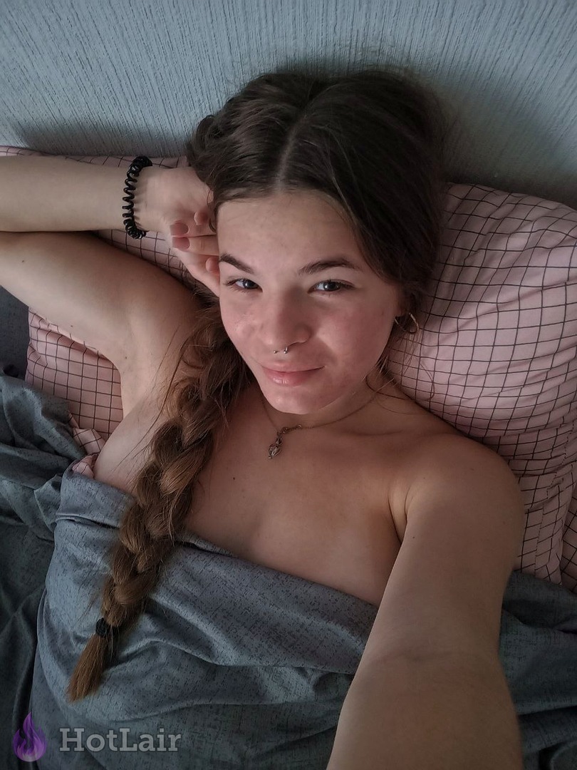 Topless brunette with braided hair lying in bed under blanket smiling at camera with seductive look
